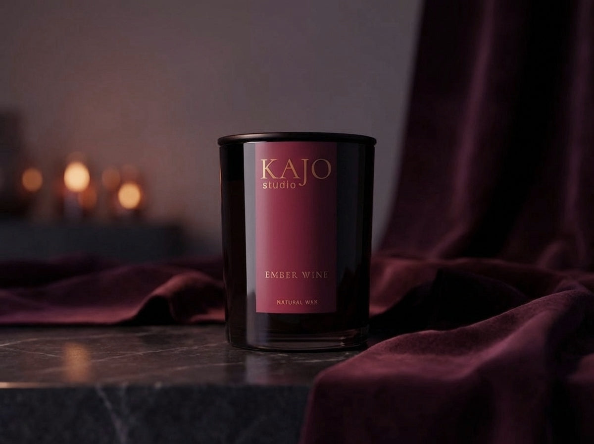 Kajo Studio candle with 'Ember Wine' scent on a dark surface with draped fabric and blurred lights in the background.