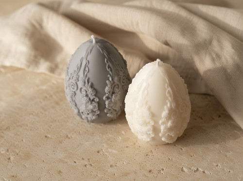Two decorative candles shaped like eggs on a textured surface with a beige fabric background